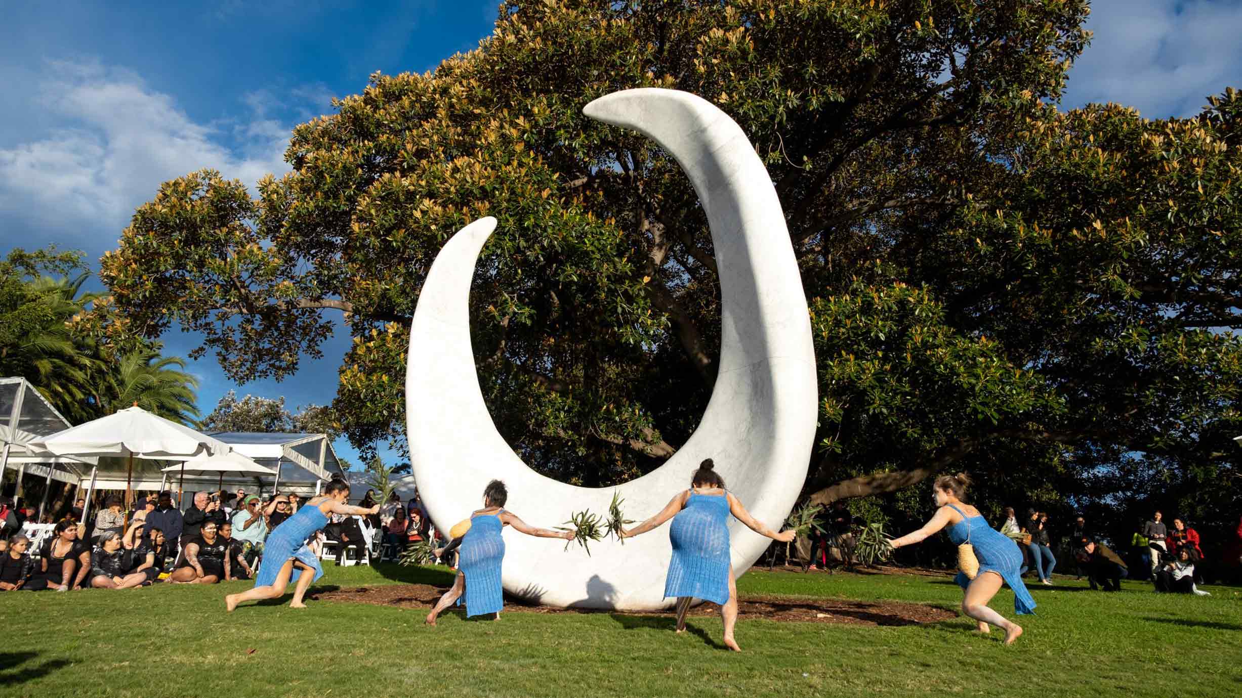 Four people in blue outfits dance in front of a large white crescent-shaped sculpture outdoors, with a crowd seated under trees in the background.